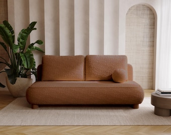 Op de afbeelding: Een bruine tweezitsbank met een afgerond ontwerp en een bijpassend rond kussen. De bank staat op een lichtgekleurd tapijt, met een grote plant in pot aan de linkerkant en een kleine bijzettafel aan de rechterkant. De achtergrond heeft neutrale gordijnen en een gebogen muur.