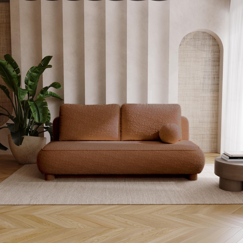 May include: A brown, two-cushion sofa with a rounded design and a matching round pillow. The sofa sits on a light-colored rug, with a large potted plant to the left and a small side table to the right. The background features neutral-toned curtains and an arched wall.