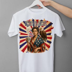 May include: White t-shirt with a graphic design. The design includes the text "SPANGLED, SAVAGE & BARELY CAFFEINATED", an illustration of a woman, an eagle, a skull, and a coffee mug with the text "1776 PROBLEMS".
