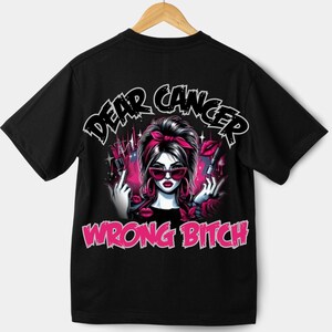 Puede incluir: Camiseta negra con un gráfico de una mujer con gafas de sol y una diadema roja, sosteniendo un lápiz labial. El texto "DEAR CANCER" está encima del gráfico, y "WRONG BITCH" debajo. El diseño es rosa y negro.