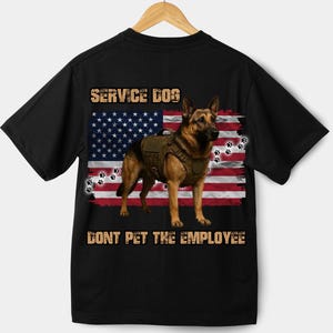 Può includere: T-shirt nera con un pastore tedesco, cane da assistenza, con un giubbotto, su sfondo di bandiera americana usurata. La maglietta include il testo "SERVICE DOG" e "DONT PET THE EMPLOYEE" in un font usurato.