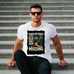 Puede incluir: Camiseta blanca con un gráfico que dice "Touch my tools & I'll file you under missing". El gráfico presenta a un mecánico sosteniendo una llave. La camiseta la lleva una persona.
