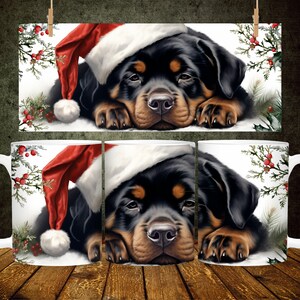 Puede incluir: Una taza de cerámica blanca con un cachorro Rottweiler negro y marrón que lleva un gorro de Papá Noel rojo. El cachorro está tumbado con la cabeza apoyada en las patas. La taza tiene un asa blanca y un fondo blanco con acebo verde y bayas rojas.