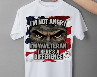 Puede incluir: Camiseta blanca con el texto "I'M NOT ANGRY I'M A VETERAN THERE'S A DIFFERENCE" y un gráfico de una cara de camuflaje con ojos intensos, sobre un fondo de la bandera estadounidense.