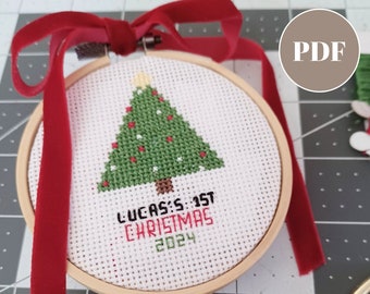 May include: A cross stitch pattern for a baby's first Christmas ornament. The pattern features a green Christmas tree with red ornaments and the text "Lucas's 1st Christmas 2024".