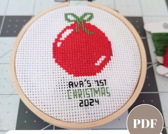 May include: A white cross stitch pattern with a red ornament and the text "AVA'S 1ST CHRISTMAS 2024". This is a PDF download for a customizable baby's first Christmas ornament.