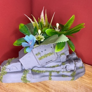 May include: A grey, 3D printed planter shaped like a Star Wars AT-AT walker with green moss and artificial plants and flowers in blue and white.