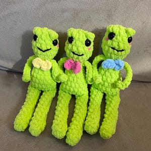 May include: Three crocheted green frog plushies with different colored bows. The frogs have black eyes and pink cheeks.