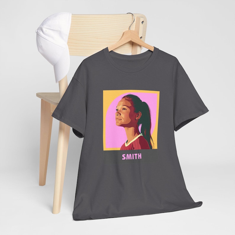May include: A charcoal gray t-shirt with a graphic of a woman with a pink background and the word "SMITH" printed below the graphic.