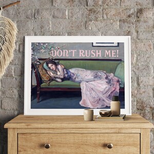 May include: A vintage-style print featuring a woman lying on a couch with the text "Don't Rush Me!" in pink lettering. The woman is wearing a flowing pink and white dress and has her head resting on a pillow. The couch is green and has a floral pattern on the back.