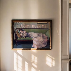 May include: A framed painting of a woman lying on a green couch. The woman is wearing a white dress and has her head resting on a pillow. The painting is titled "Don't Rush Me!"