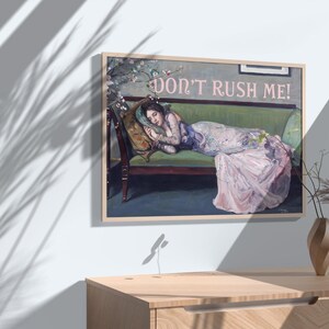 May include: A framed painting of a woman lying on a green couch with the text "Don't Rush Me!" in pink letters. The woman is wearing a pink dress and has her head resting on a pillow. The painting is set against a white wall with a shadow of a plant.