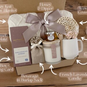 May include: A gift box filled with cozy items, including a knit throw blanket, gourmet chocolate, a ceramic mug, a woven coaster, a French lavender candle, honey and a honey dipper, and a beverage in a burlap sack.