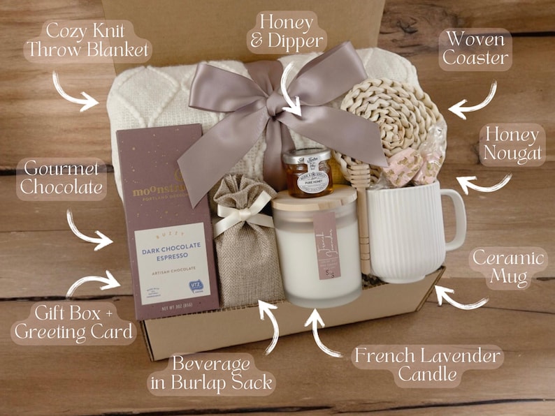 May include: A gift box filled with cozy items, including a knit throw blanket, gourmet chocolate, a ceramic mug, a woven coaster, a French lavender candle, honey and a honey dipper, and a beverage in a burlap sack.