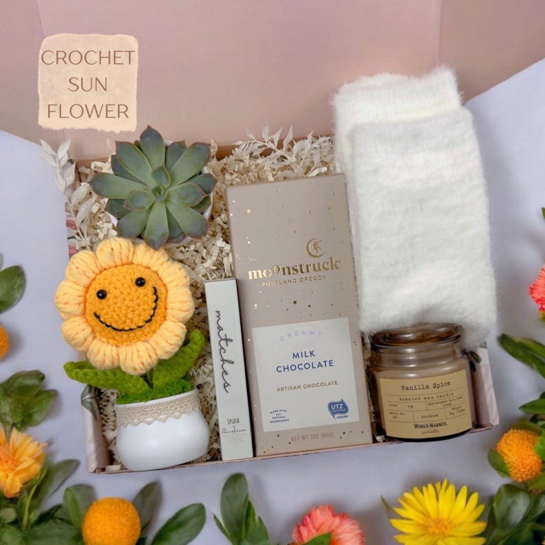 K&ouml;nnte beinhalten: Eine Geschenkbox mit einer gelben H&auml;kel-Sonnenblume in einem wei&szlig;en Topf, einer gr&uuml;nen Sukkulente, einer Streichholzschachtel, einer Moonstruck-Milchschokoladentafel, einer kleinen Vanilla Spice-Kerze und flauschigen wei&szlig;en Socken.