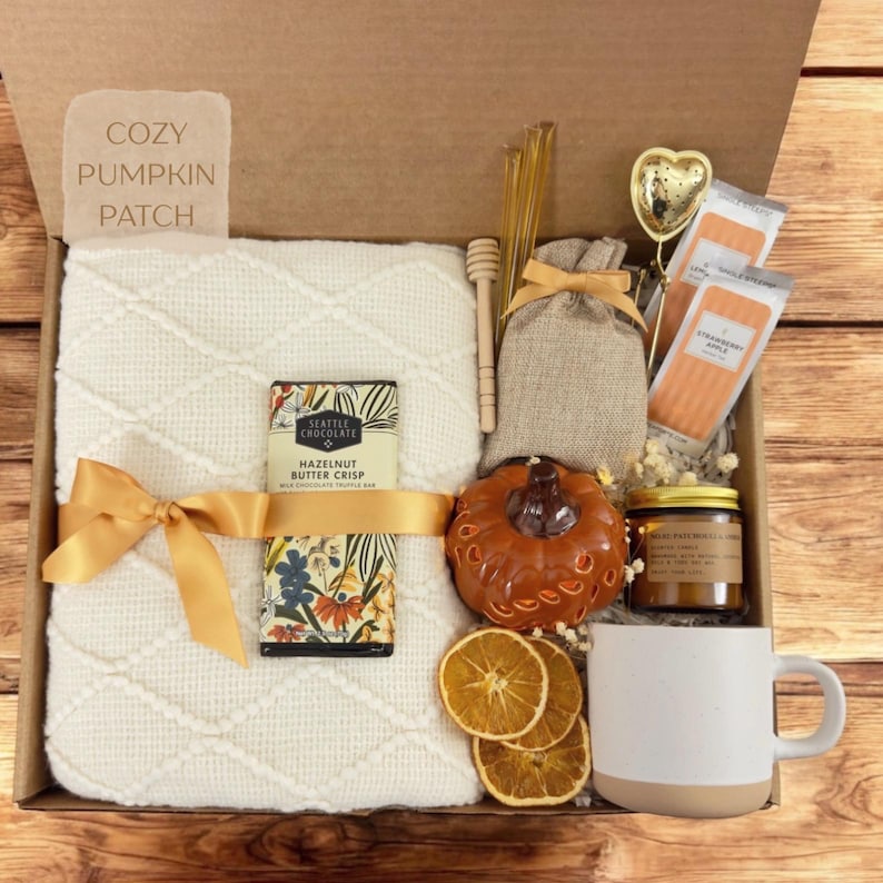 May include: A gift box labeled Cozy Pumpkin Patch containing a cream-colored textured throw blanket with a gold ribbon, a Seattle Chocolate hazelnut butter crisp bar, a burlap pouch, a ceramic pumpkin, a candle, tea, and a white ceramic mug.