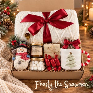 May include: A gift box containing a white cable-knit blanket tied with a red ribbon, a snowman figurine, a jar of vanilla lavender candle, a jar of marshmallows, a bar of soap, red-wrapped candies, and a white mug with a Christmas tree design.