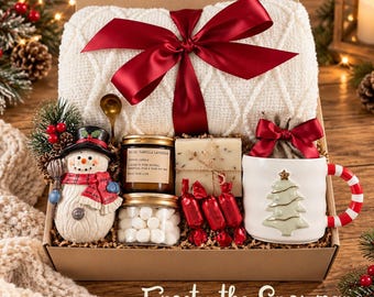 May include: A gift box containing a white cable-knit blanket tied with a red ribbon, a snowman figurine, a jar of vanilla lavender candle, a jar of marshmallows, a bar of soap, red-wrapped sweets, and a white mug with a Christmas tree design.
