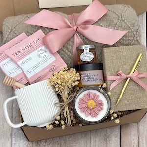 May include: A gift box filled with self-care items, including tea, honey, body butter, a candle, a journal, and a pen. The box is decorated with a pink ribbon and a blanket.