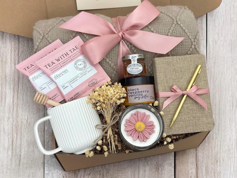 May include: A gift box filled with self-care items, including tea, honey, body butter, a candle, a journal, and a pen. The box is decorated with a pink ribbon and a blanket.