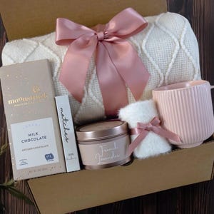 K&ouml;nnte beinhalten: Eine Geschenkbox gef&uuml;llt mit gem&uuml;tlichen Artikeln, darunter eine wei&szlig;e Decke mit einer rosa Schleife, eine rosa Tasse, eine Kerze, eine Tafel Milchschokolade und ein kleines, wei&szlig;es, flauschiges Teil. Die Box ist aus braunem Karton und hat einen nat&uuml;rlichen Holz Hintergrund.