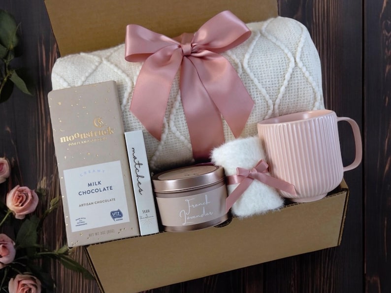 K&ouml;nnte beinhalten: Eine Geschenkbox gef&uuml;llt mit gem&uuml;tlichen Artikeln, darunter eine wei&szlig;e Decke mit einer rosa Schleife, eine rosa Tasse, eine Kerze, eine Tafel Milchschokolade und ein kleines, wei&szlig;es, flauschiges Teil. Die Box ist aus braunem Karton und hat einen nat&uuml;rlichen Holz Hintergrund.