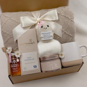 May include: A gift basket filled with a cozy blanket, a mug, a jar of honey, a bar of chocolate, a small plush potato toy, and a candle. The basket is wrapped in a beige and white patterned paper and tied with a white satin ribbon.