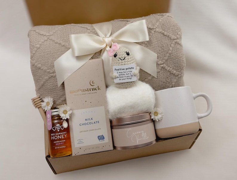 May include: A gift basket filled with a cozy blanket, a mug, a jar of honey, a bar of chocolate, a small plush potato toy, and a candle. The basket is wrapped in a beige and white patterned paper and tied with a white satin ribbon.
