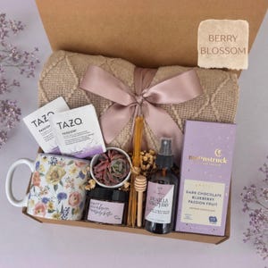 May include: A gift box containing a beige textured throw blanket with a mauve ribbon, a floral ceramic mug, two Tazo Passion tea bags, a small succulent, a jar of berry blossom body butter, a wooden honey dipper, a vanilla orchid spray, and a Moonstruck dark chocolate bar.