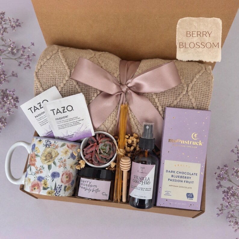 May include: A gift box containing a beige textured throw blanket with a mauve ribbon, a floral ceramic mug, two Tazo Passion tea bags, a small succulent, a jar of berry blossom body butter, a wooden honey dipper, a vanilla orchid spray, and a Moonstruck dark chocolate bar.