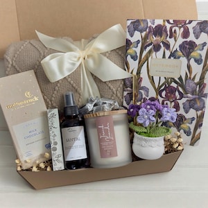 May include: A gift basket filled with items for relaxation and self-care. It includes a throw blanket, a candle, a bottle of essential oil, a bar of chocolate, a notebook, and a small potted plant.