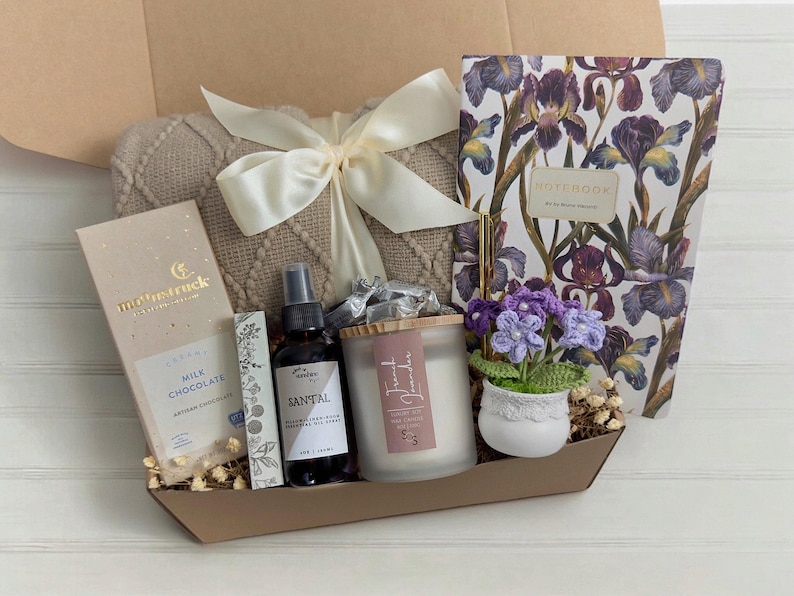 May include: A gift basket filled with items for relaxation and self-care. It includes a throw blanket, a candle, a bottle of essential oil, a bar of chocolate, a notebook, and a small potted plant.