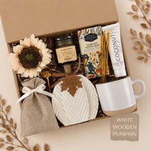 May include: A gift box containing a cream-colored sunflower, a small candle labeled Harvest Leaves, a Seattle Chocolate bar, a tube of Coconut hand cream, a gold spoon, a burlap pouch with a white ribbon, and a white ceramic mug.