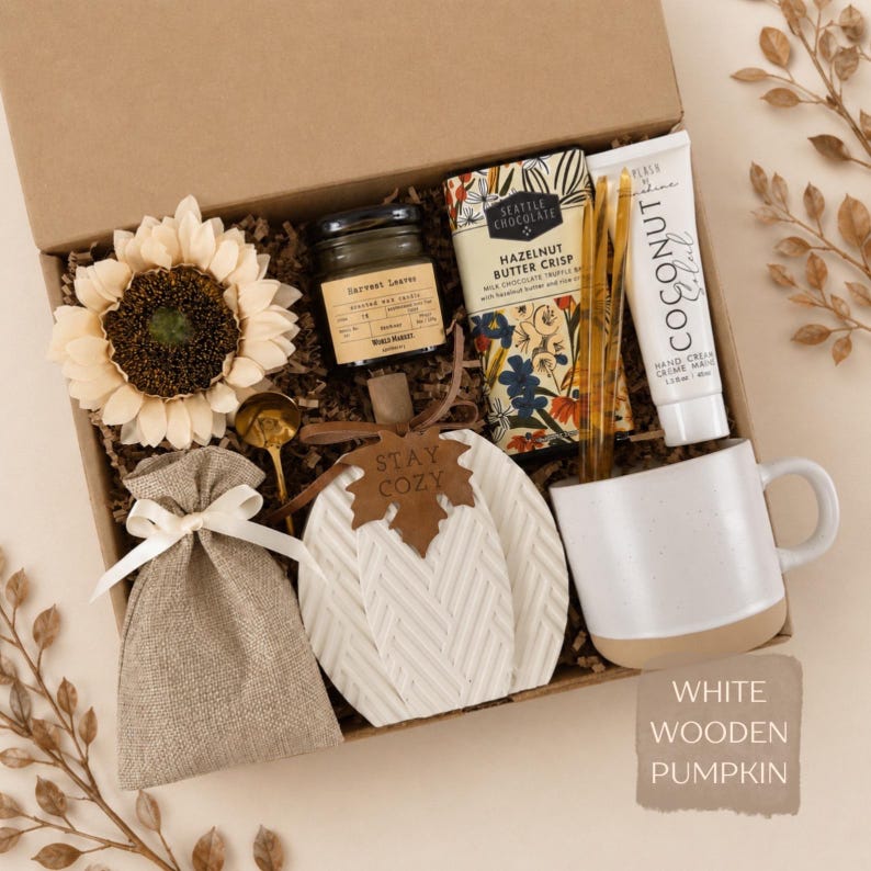 May include: A gift box containing a cream-colored sunflower, a small candle labeled Harvest Leaves, a Seattle Chocolate bar, a tube of Coconut hand cream, a gold spoon, a burlap pouch with a white ribbon, and a white ceramic mug.