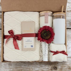 May include: A gift box filled with self-care items, including a white throw blanket, a candle, a glass tumbler, a bar of chocolate, a tube of hand cream, and a washcloth. The box is decorated with a red ribbon and a red sunflower.