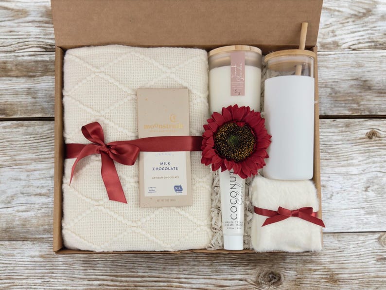 May include: A gift box filled with self-care items, including a white throw blanket, a candle, a glass tumbler, a bar of chocolate, a tube of hand cream, and a washcloth. The box is decorated with a red ribbon and a red sunflower.