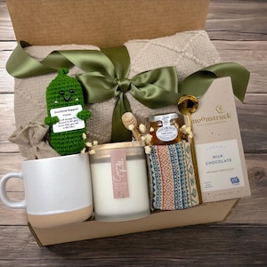 May include: A gift box filled with a variety of items, including a crocheted pickle toy, a candle, a jar of honey, a mug, a blanket, and a bar of milk chocolate. The box is decorated with a green ribbon and a tag that reads "Emotional Support Pickle".