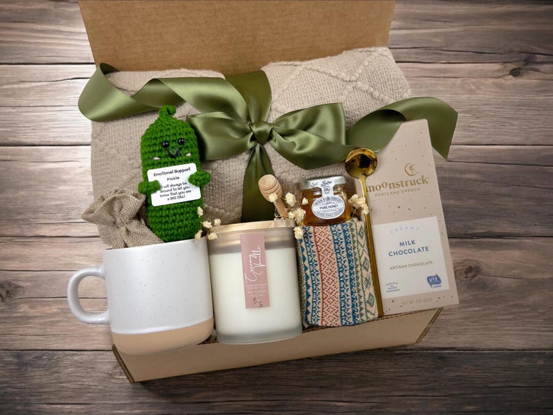 May include: A gift box filled with a variety of items, including a crocheted pickle toy, a candle, a jar of honey, a mug, a blanket, and a bar of milk chocolate. The box is decorated with a green ribbon and a tag that reads "Emotional Support Pickle".