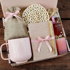 May include: A pink and brown gift box filled with a pink mug, a notebook, a woven coaster, a small bag, a honey jar, and a pink candle. The box is decorated with pink ribbon and a floral pattern.