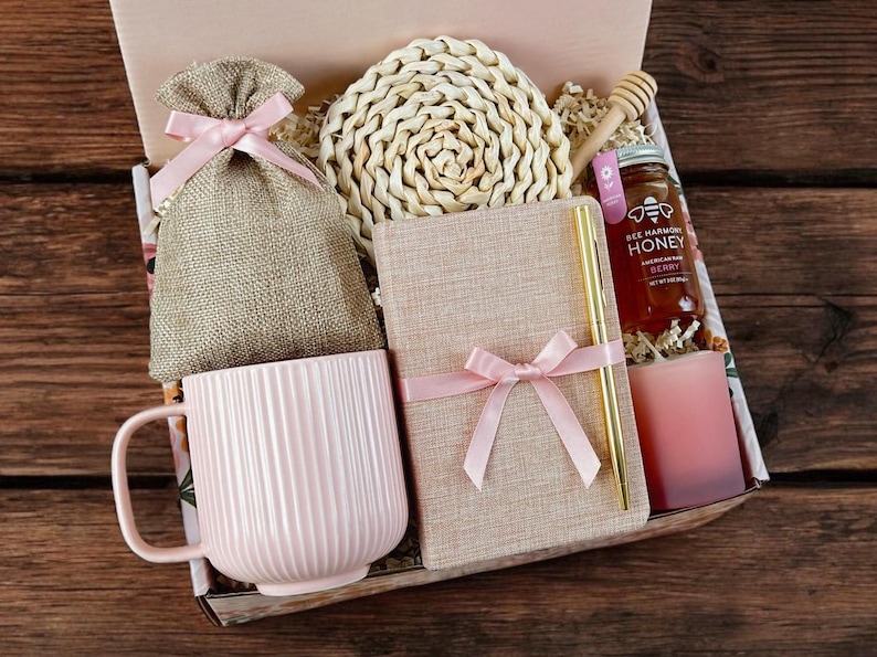 May include: A pink and brown gift box filled with a pink mug, a notebook, a woven coaster, a small bag, a honey jar, and a pink candle. The box is decorated with pink ribbon and a floral pattern.