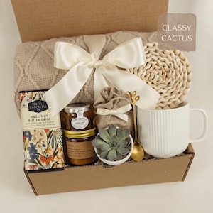 May include: A gift box containing a beige knit blanket tied with a white satin ribbon, a Seattle Chocolate bar, a jar of honey, a small burlap bag, a green succulent in a white pot, a gold spoon, and a round woven coaster next to a white mug.