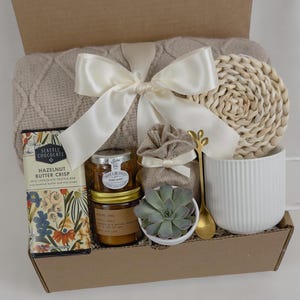 May include: A gift box filled with cozy items, including a beige throw blanket with a white satin ribbon, a white mug, a small potted succulent, a jar of candle, a chocolate bar, and a woven coaster.
