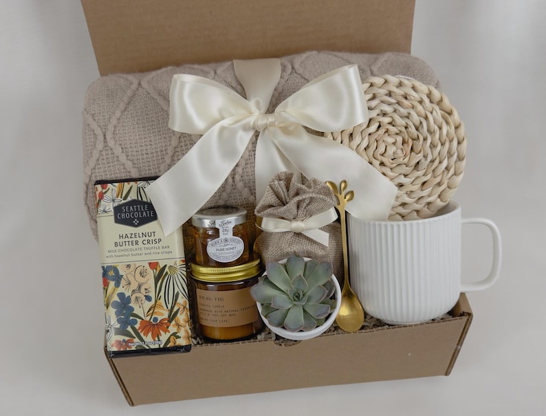 May include: A gift box filled with cozy items, including a beige throw blanket with a white satin ribbon, a white mug, a small potted succulent, a jar of candle, a chocolate bar, and a woven coaster.