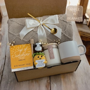 K&ouml;nnte beinhalten: Eine Geschenkbox mit einer beigen, strukturierten Decke, die mit einem wei&szlig;en Band und einem goldenen L&ouml;ffel verziert ist, Big Heart Tee, einem kleinen Honigglas, einer geh&auml;kelten Ananas mit Schild Fine-Apple It's fine, einer Kerze und einem Becher.