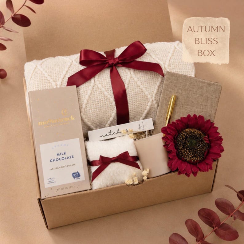 May include: A gift box labeled AUTUMN BLISS BOX containing a white cable-knit blanket tied with a burgundy ribbon, a milk chocolate bar, a small white fuzzy pillow with a burgundy ribbon, a beige candle, a notebook, a gold pen, and a dark red flower.