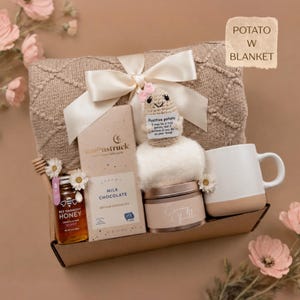 K&ouml;nnte beinhalten: Eine Geschenkbox mit einer beigen Strickdecke, die mit einem wei&szlig;en Band gebunden ist, einer kleinen geh&auml;kelten Kartoffelfigur mit einem Schild mit der Aufschrift Positive potato, einer Moonstruck-Milchschokolade, Bee Harmony-Honig und einem wei&szlig;en Becher.