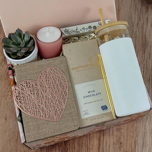 May include: A gift box filled with items including a small potted succulent, a pink candle, a bar of milk chocolate, a notebook with a rose gold heart design, and a white tumbler with a bamboo lid and straw.
