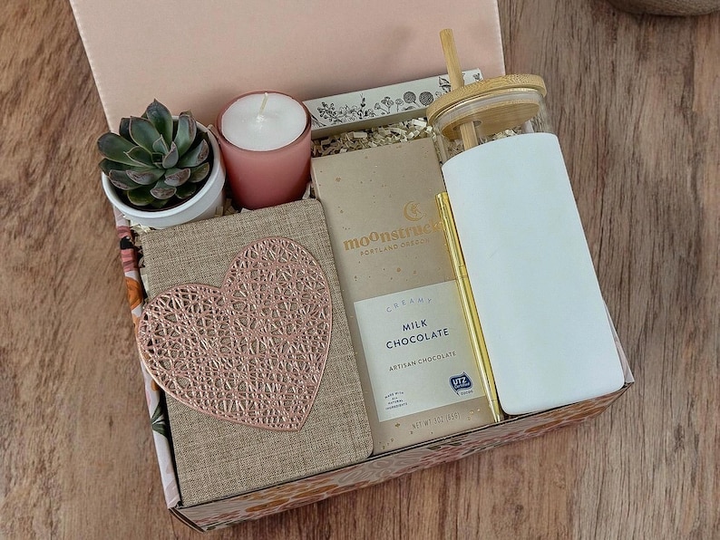 May include: A gift box filled with items including a small potted succulent, a pink candle, a bar of milk chocolate, a notebook with a rose gold heart design, and a white tumbler with a bamboo lid and straw.