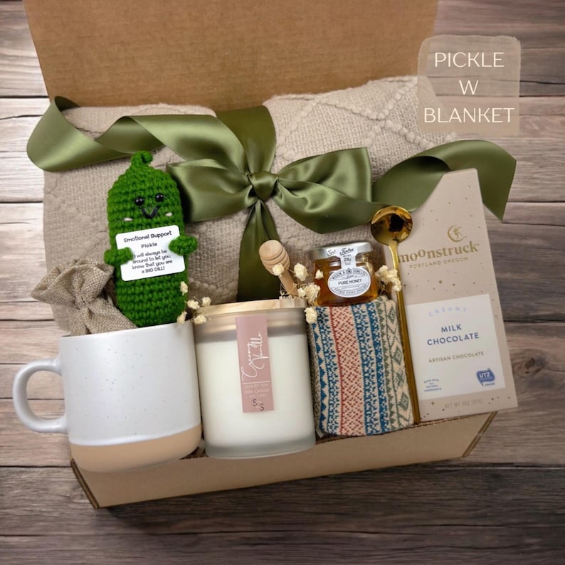 K&ouml;nnte beinhalten: Eine Geschenkbox mit einer beigen, strukturierten Decke mit gr&uuml;nem Band, einem geh&auml;kelten gr&uuml;nen Gurkenspielzeug mit Schild, einer wei&szlig;en Keramiktasse, einer Creamy Vanilla Kerze, einem Honigglas mit L&ouml;ffel, einem gemusterten Stoff und einer Milchschokolade.