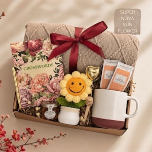 K&ouml;nnte beinhalten: Eine Geschenkbox mit einer beigen Strickdecke, die mit einem weinroten Band gebunden ist, einem floralen Kreuzwortr&auml;tselbuch, einem goldenen Stift, einem kleinen Honigglas, einer geh&auml;kelten Sonnenblume, einem herzf&ouml;rmigen Tee-Ei, zwei Teepackungen und einer Tasse.